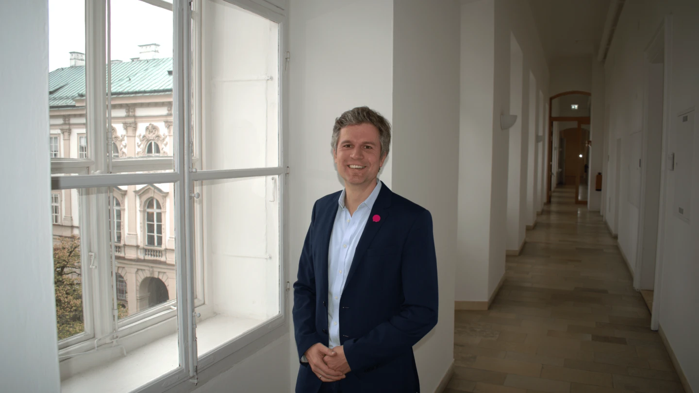 Mann im dunkelblauen Anzug lächelt vor einem Fenster mit Blick auf ein Palais in Wien.