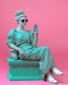 Statue einer Frau mit Sonnenbrille, die auf einem Sockel sitzt, pinke Sonnenbrillen trägt und in ihr Handy schaut.