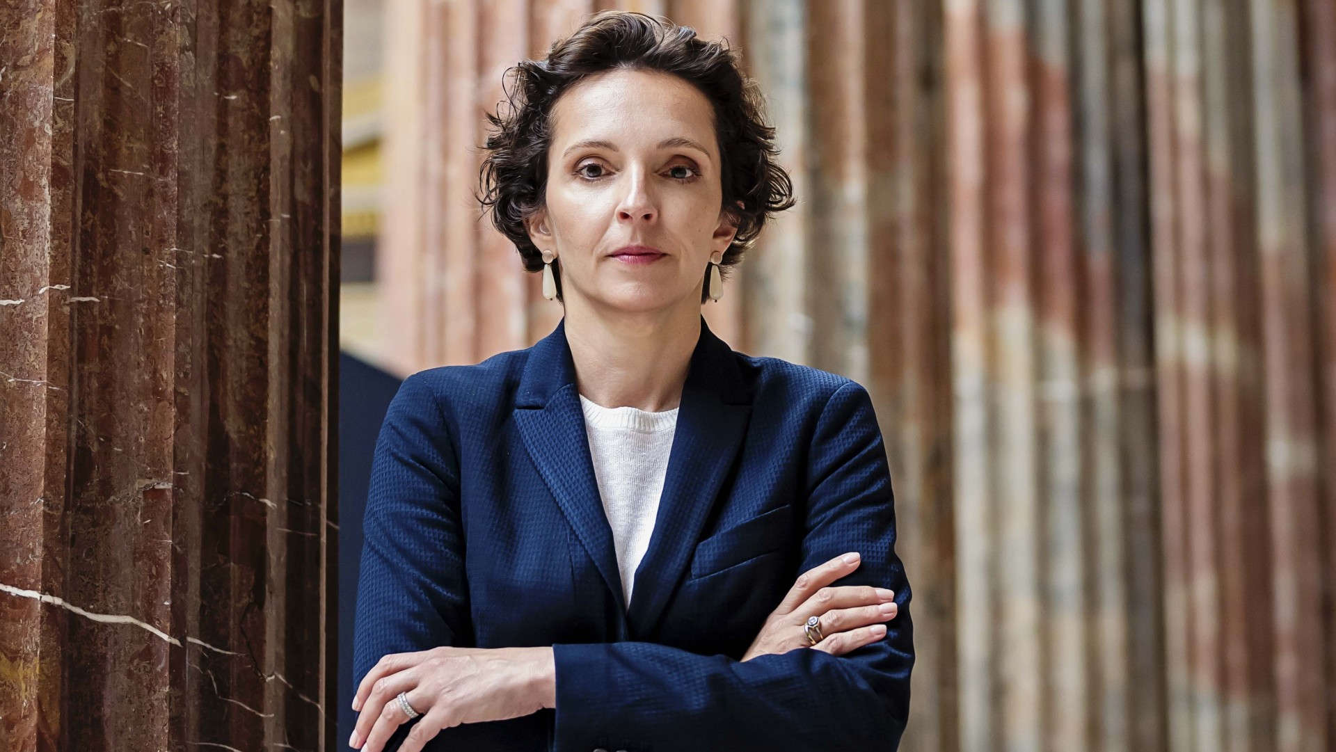 Martina Künsberg Sarre steht lächelnd vor einer Säule in formeller Kleidung.