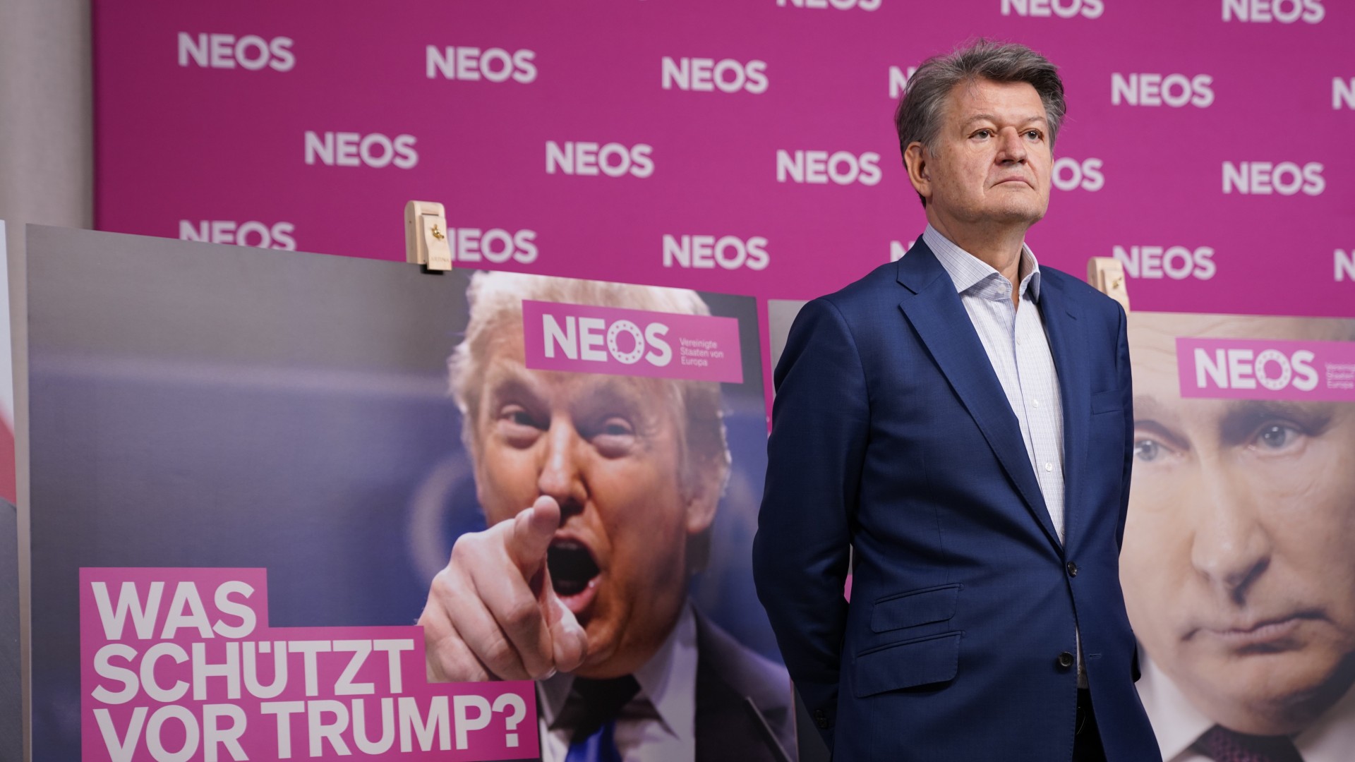 NEOS Helmut Brandstätter.
Mann in Anzug steht vor einem Plakat von Trump