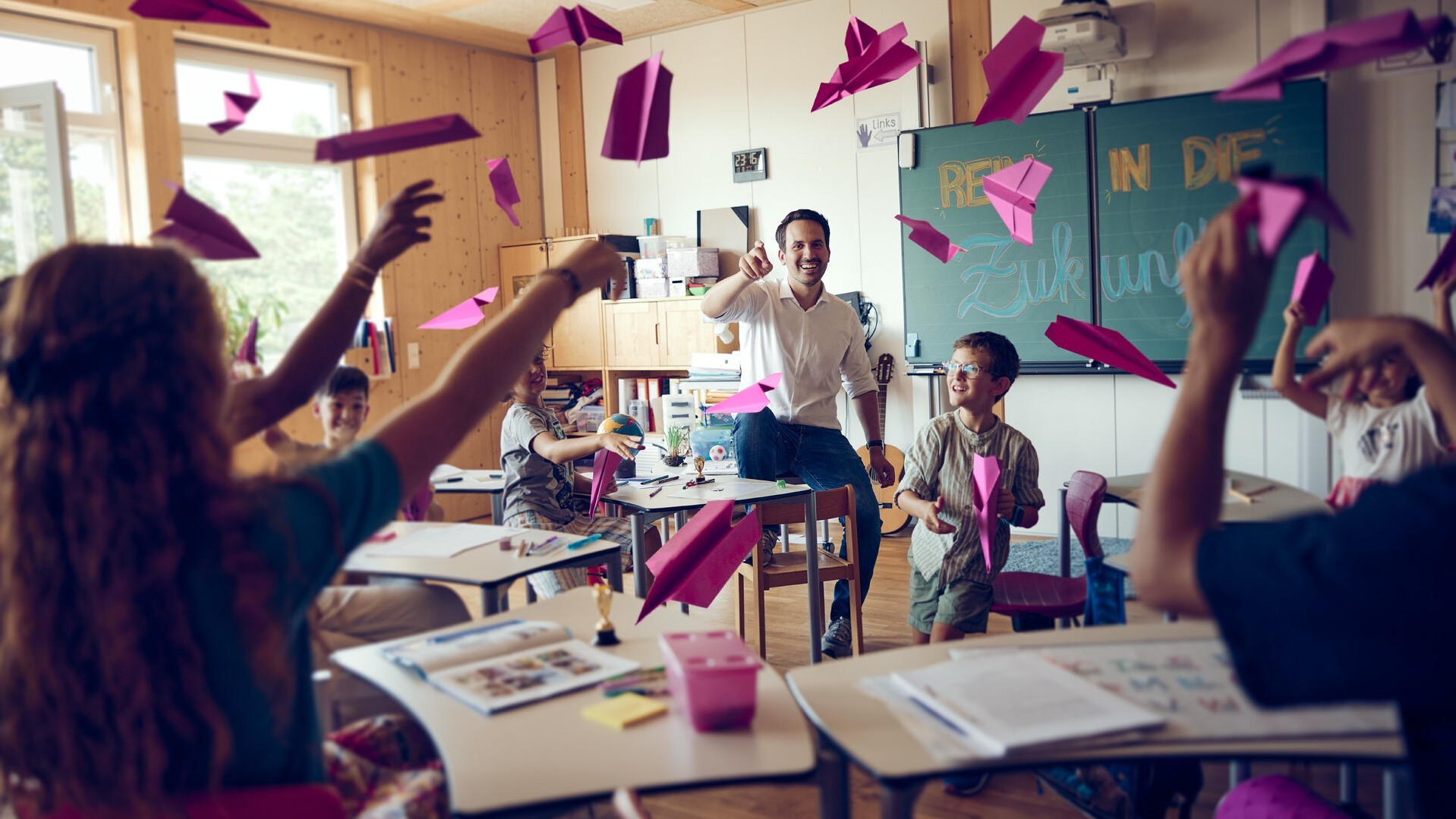 © Konstantin Reyer Christoph Wiederkehr wirft mit Schulkindern in einem Klassenzimmer pinke Papierflieger (© Konstantin Reyer)