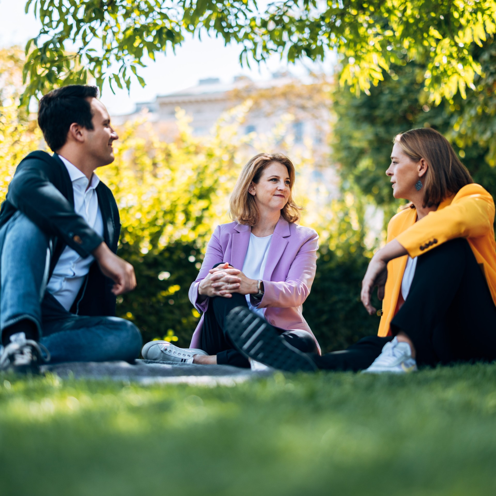 Beate Meinl-Reisinger, Christoph Wiederkehr und Andrea Klambauer sitzen gemeinsam auf einer Wiese und plaudern entspannt, während sie über politische Arbeit und Reformen sprechen.